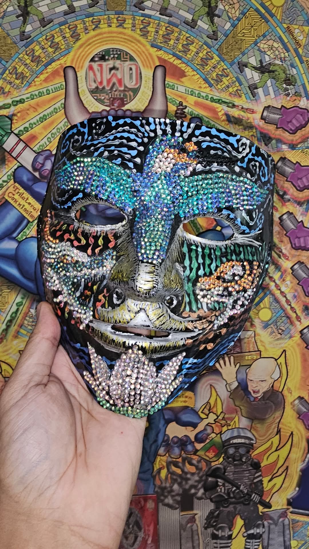 A hand holds a black mask encrusted with holographic rhinestones in a bird shape across the forehead and nose, with painted flames and a rhinestone lotus at the chin, in front of a golden conspiracy-poster background with an NWO emblem and binary rings.