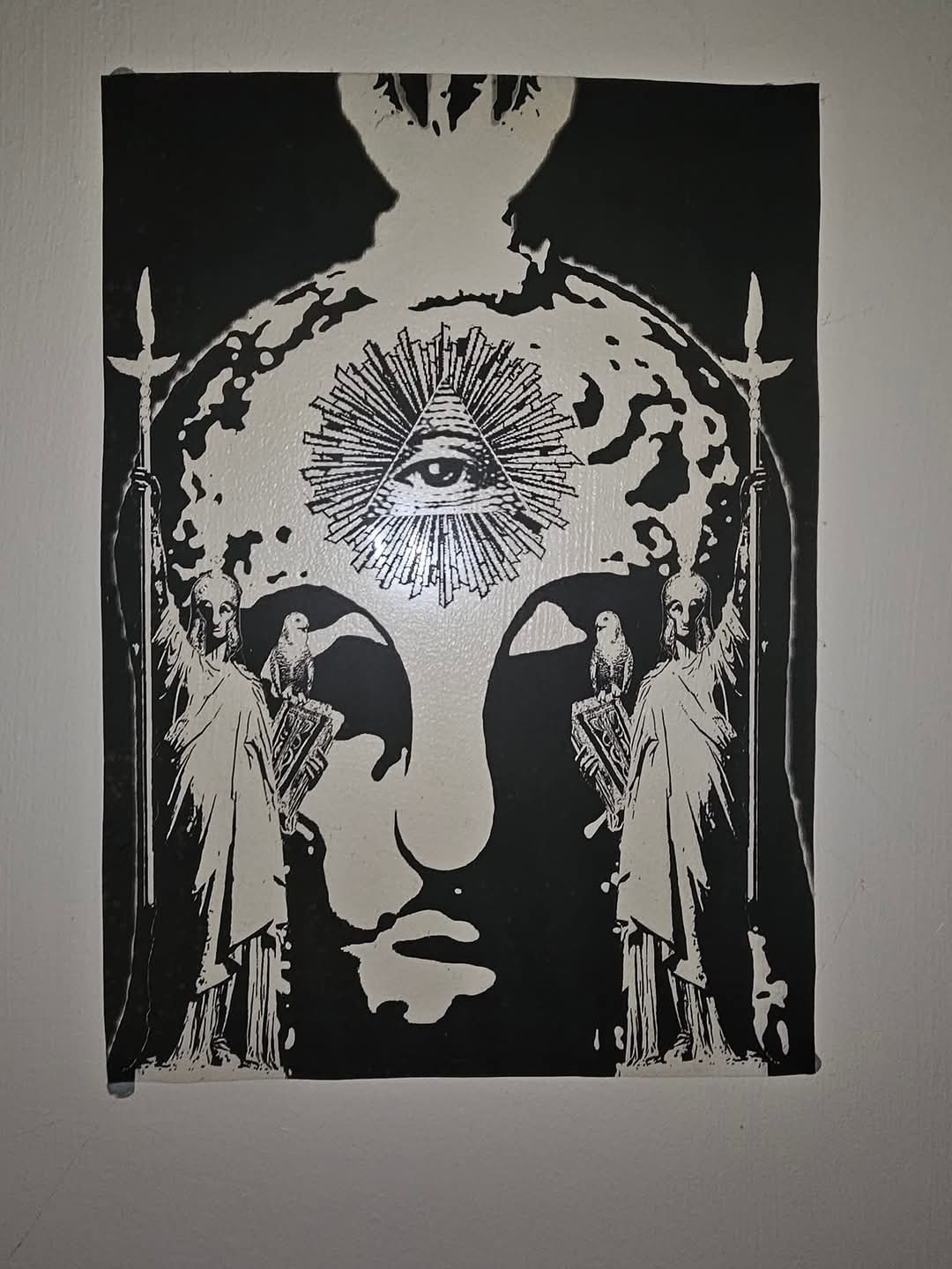 High-contrast black and white print of two mirrored robed classical figures holding a spear and a book topped by an owl, whose negative space forms a large hooded face with an eye inside a pyramid at the forehead.