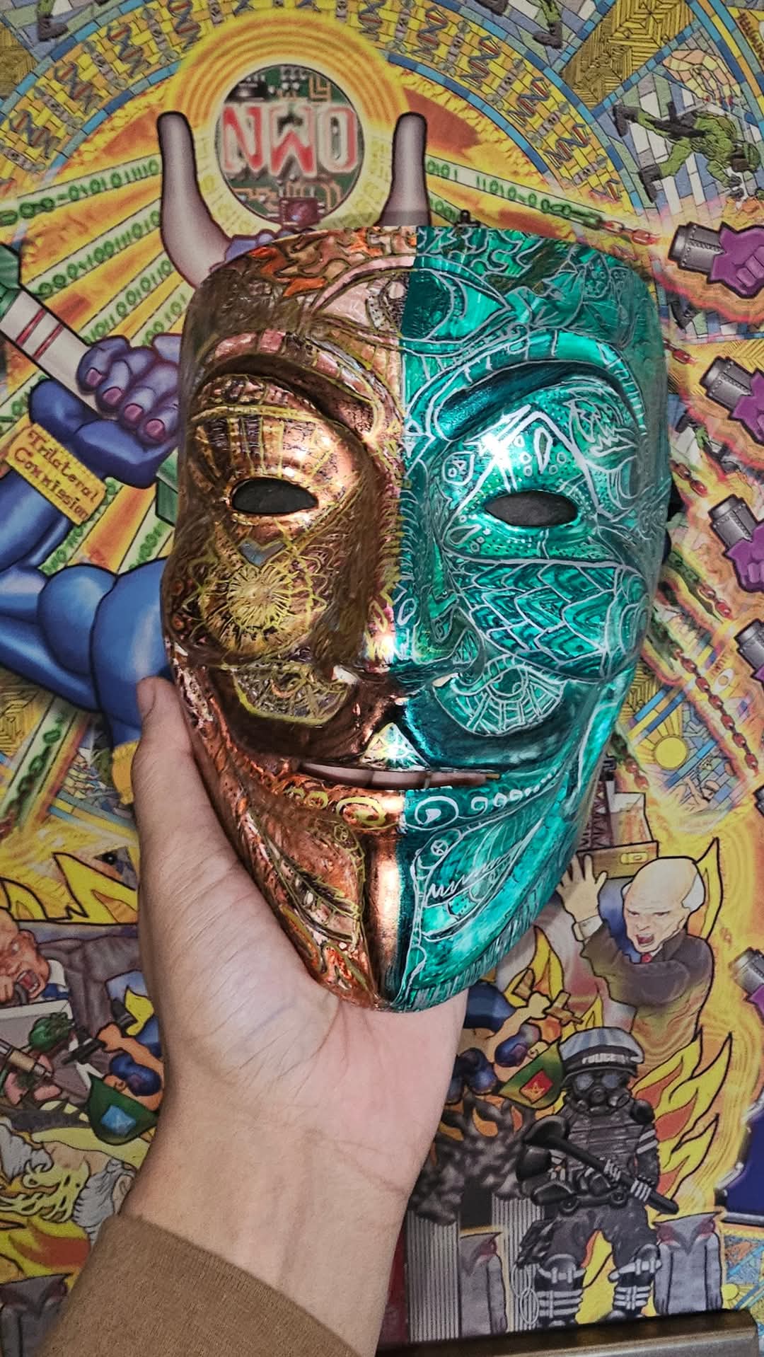 A hand holds a Guy Fawkes mask painted in two halves - copper and gold mandala patterns on one side, emerald green and teal scales on the other - in front of a bright conspiracy-poster background with an NWO emblem, binary rings and a riot officer.